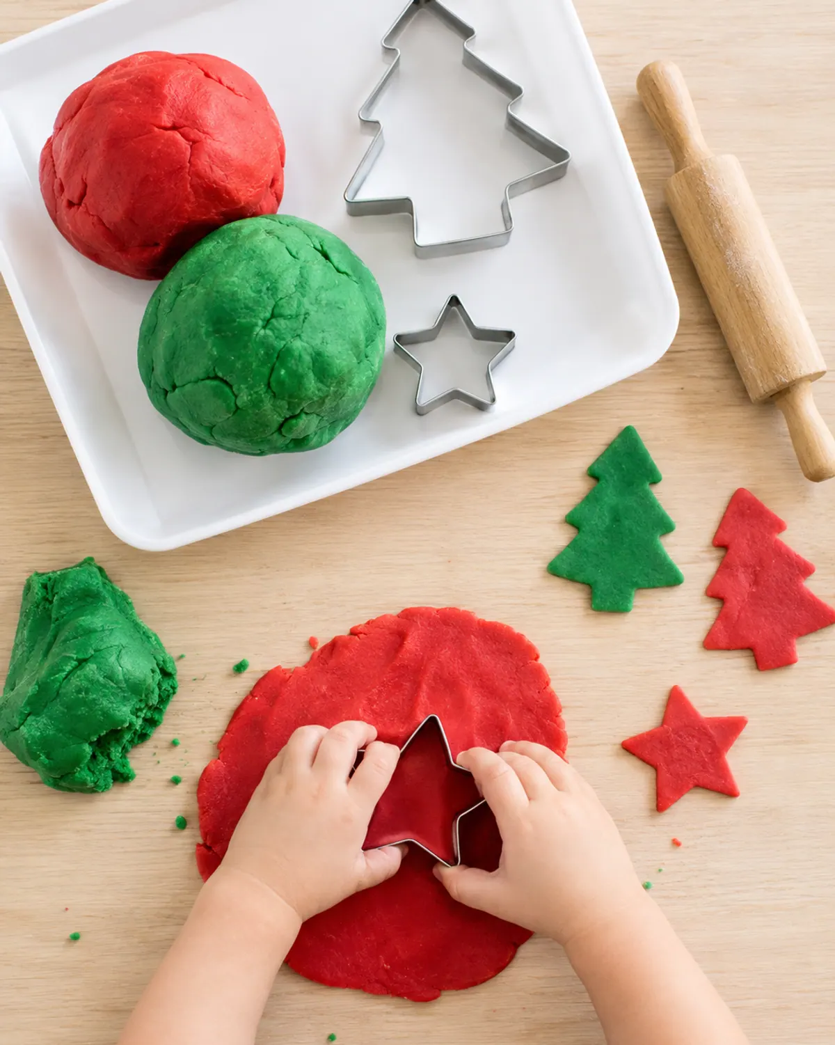 A tray of red and green playdough with Christmas tree and star cookie cutters and a small wooden rolling pin, a toddler's hands pressing a cutter