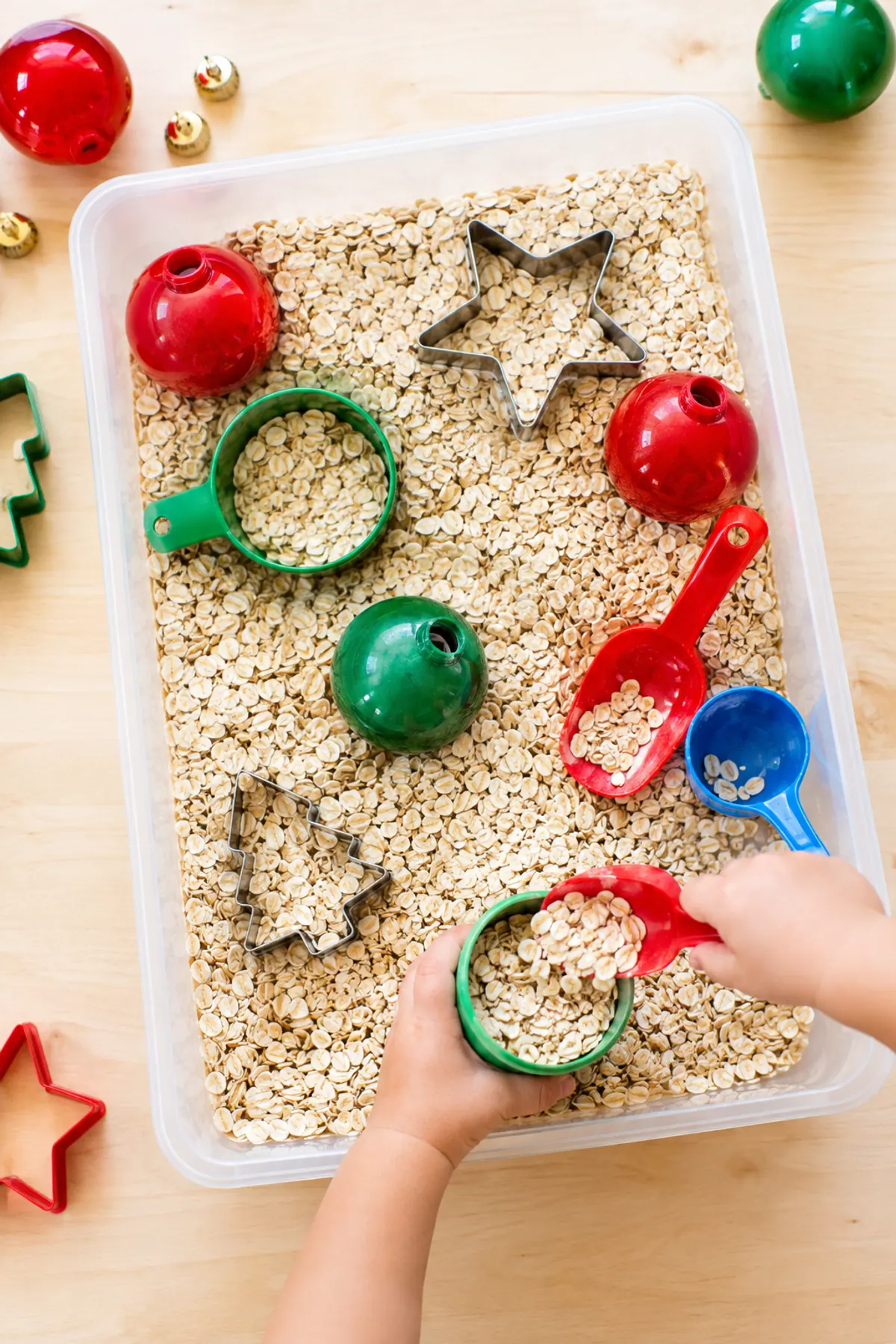 Christmas sensory bin for toddlers: a wide bin of oat snow with big cookie cutters, chunky scoops and large plastic ornaments, a toddler's small hands scooping