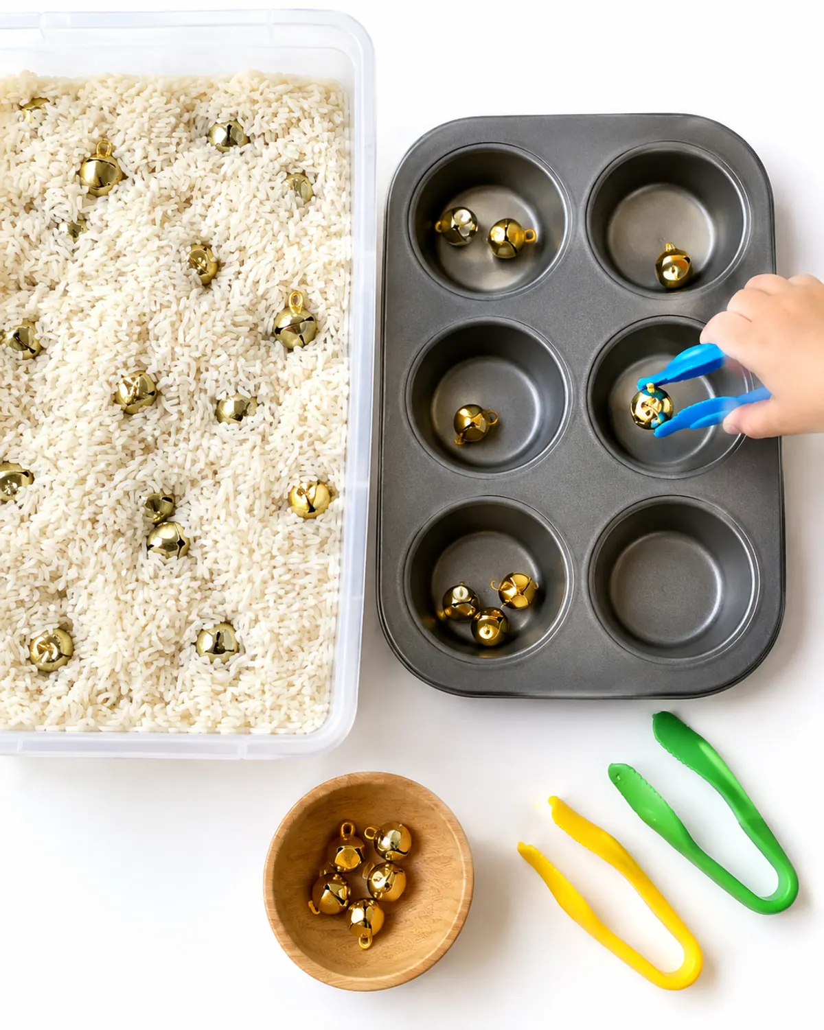 Preschool Christmas sensory bin: white rice with small gold jingle bells, a muffin tin and kid tongs, a preschooler's hand picking up a bell
