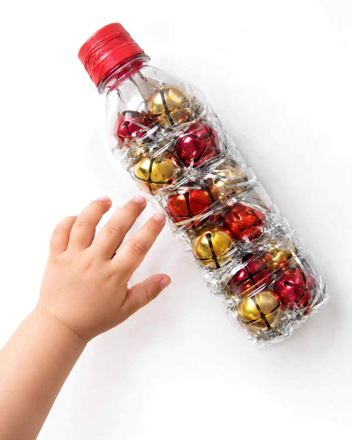 A clear plastic sensory bottle filled with gold and red jingle bells and a little tinsel, cap taped shut, lying on a play mat