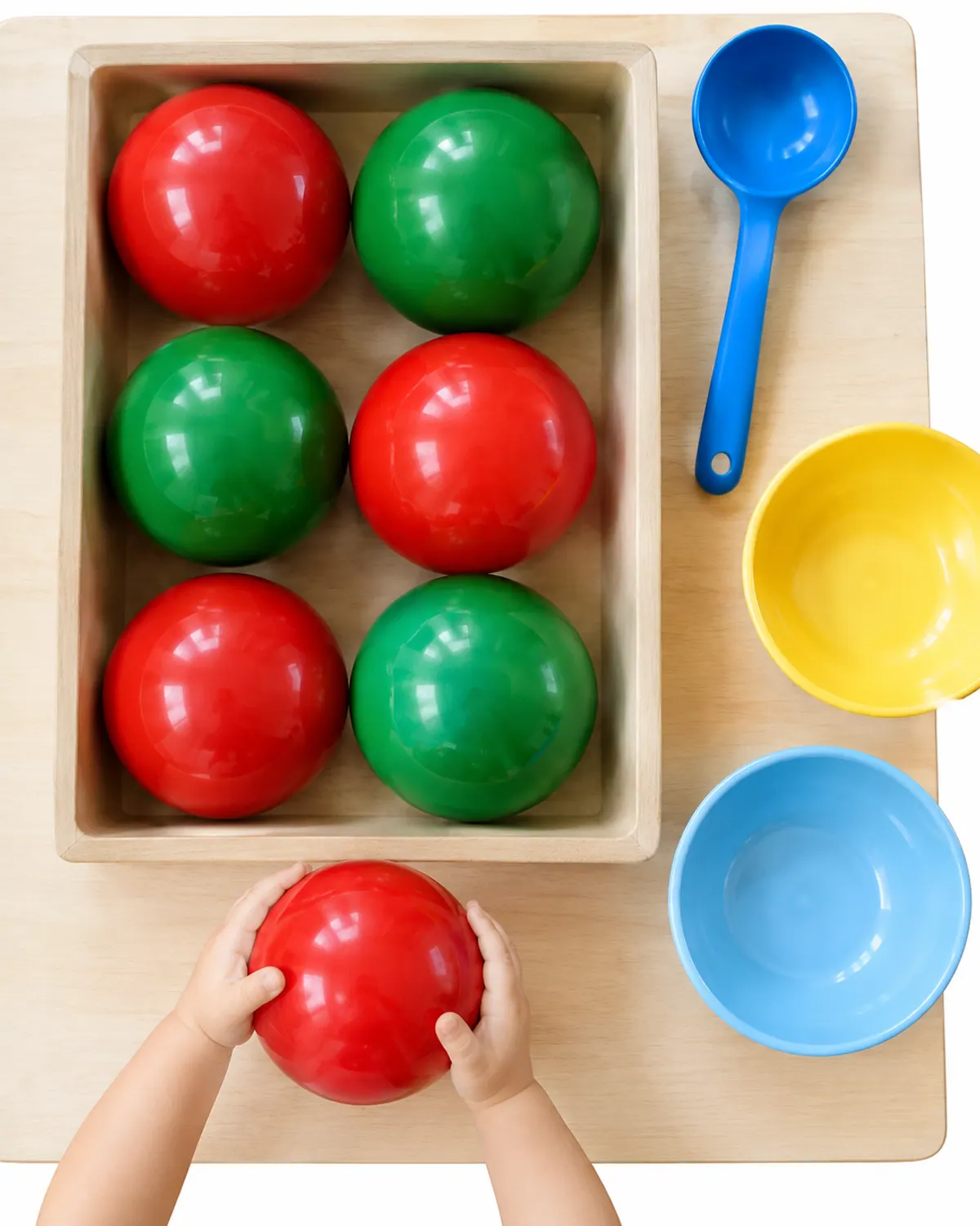 Large red and green shatterproof plastic ornaments in a wide bin with a big ladle and two bowls for sorting, on a play mat