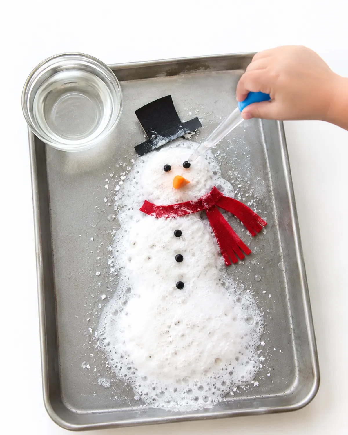 A baking soda snowman fizzing on a tray as a preschooler's hand squeezes vinegar from a dropper, a paper hat and scarf sliding off