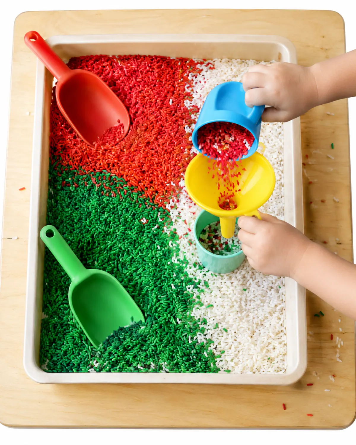 Christmas sensory bin of red, green and white dyed rice with big plastic scoops and a funnel, a toddler's hands pouring rice into a cup