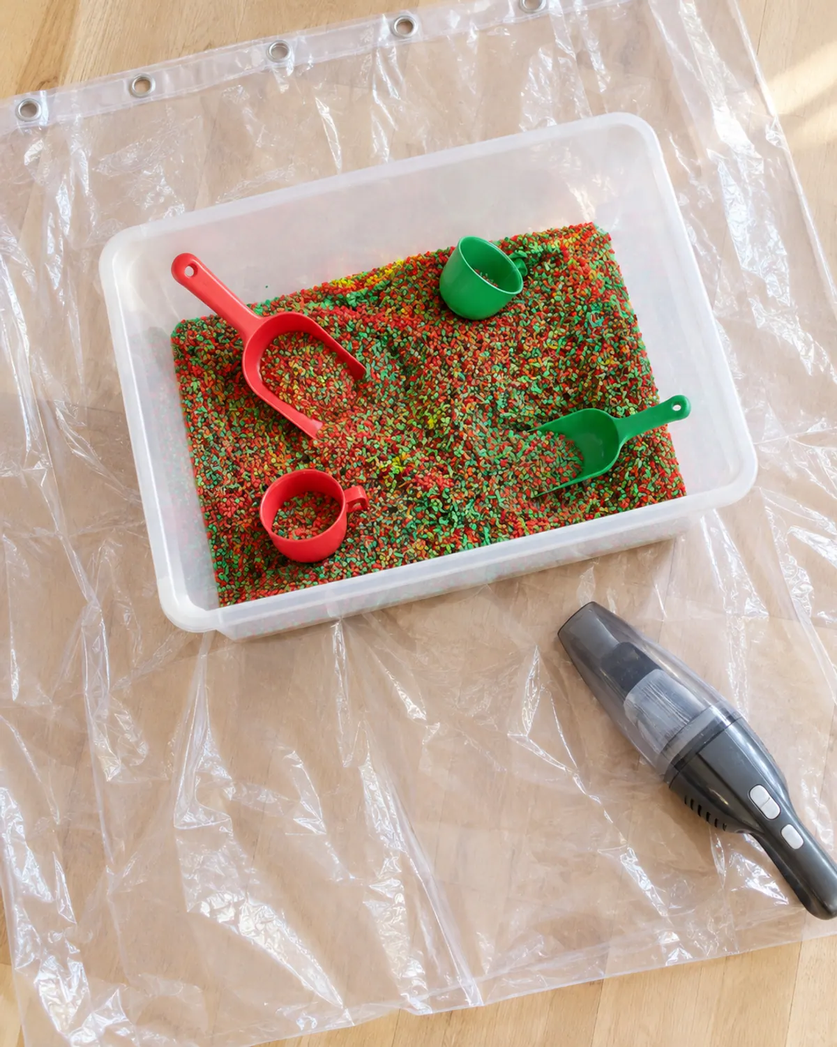 A Christmas sensory bin set on an old shower curtain on a kitchen floor, with a wide shallow bin, scoops and a handheld vacuum nearby