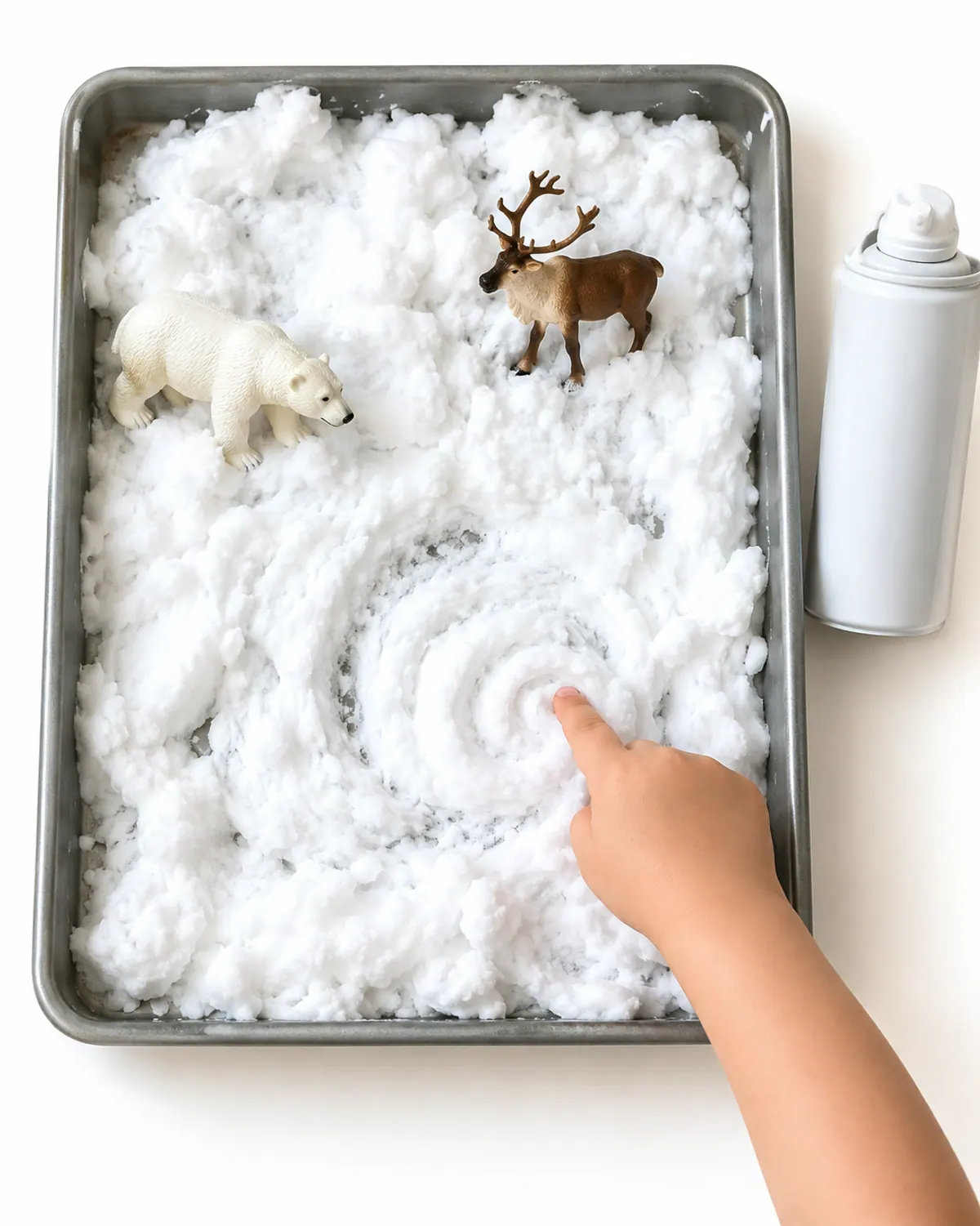 A tray of white shaving cream snow with plastic polar bear and reindeer toys, a preschooler's hand swirling the foam