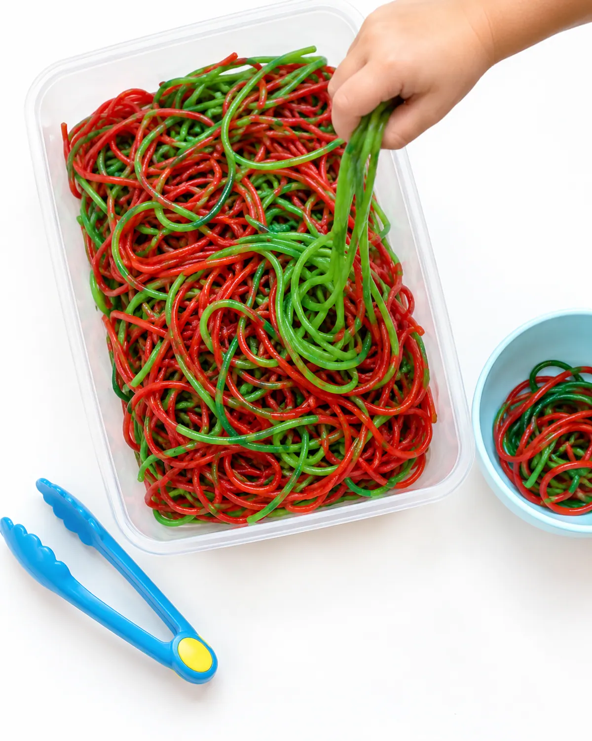 A sensory bin of cooked red and green dyed spaghetti noodles with chunky toddler tongs, a toddler's small hand pulling a strand