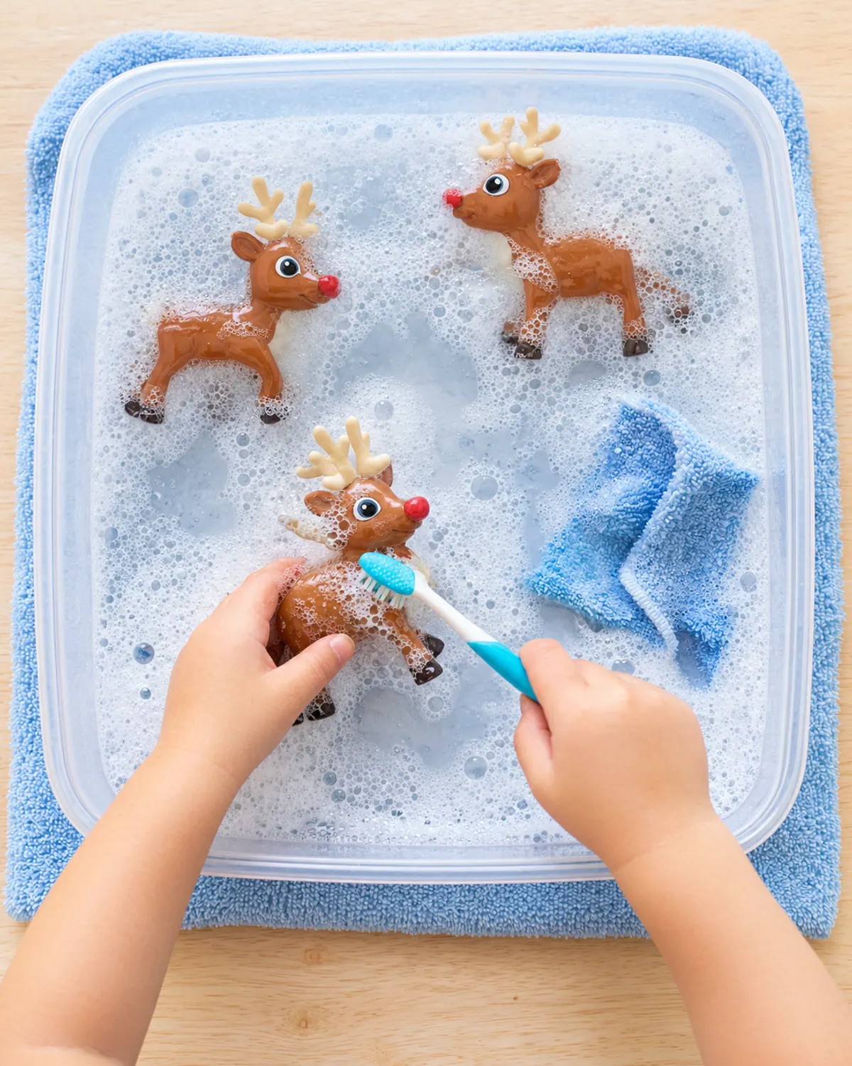 A shallow bin of soapy water with large plastic toy reindeer, a toothbrush and a washcloth, a toddler's small hand scrubbing a reindeer