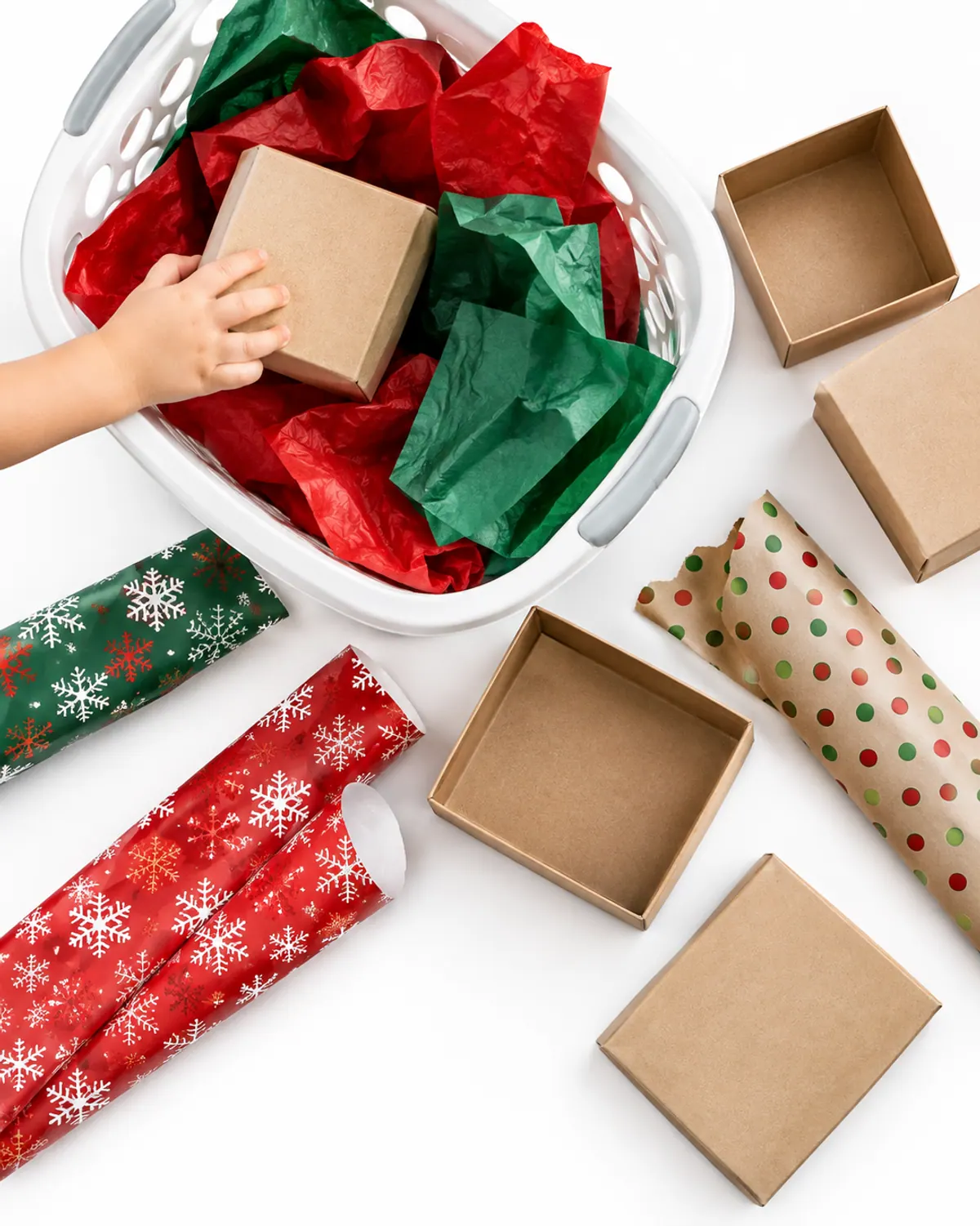 A laundry basket filled with crinkled red and green tissue paper, wrapping paper scraps and empty kraft gift boxes, a toddler's hand pulling out a box