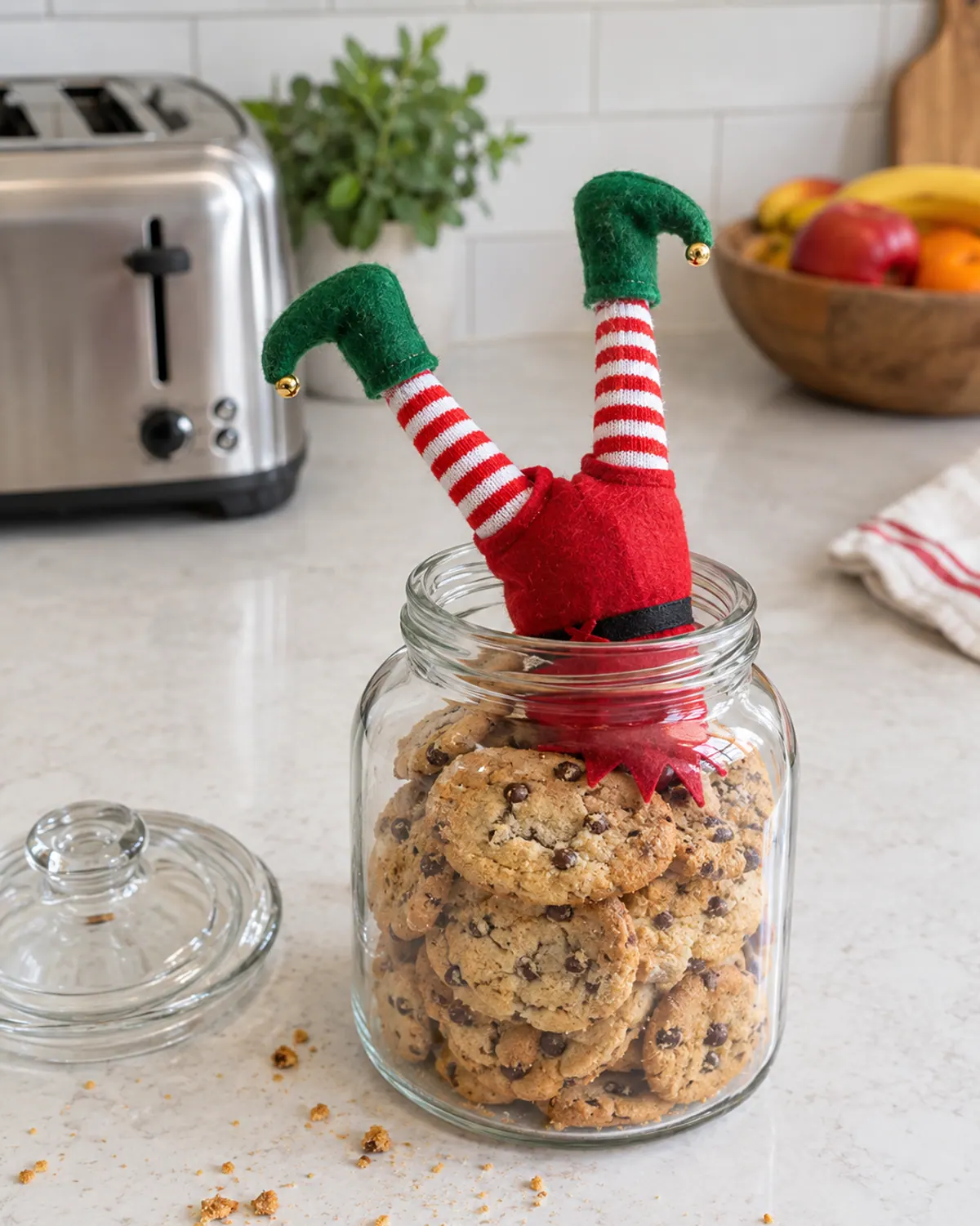 A felt elf doll stuck upside down in a glass jar of chocolate chip cookies with only its striped legs and green boots poking out
