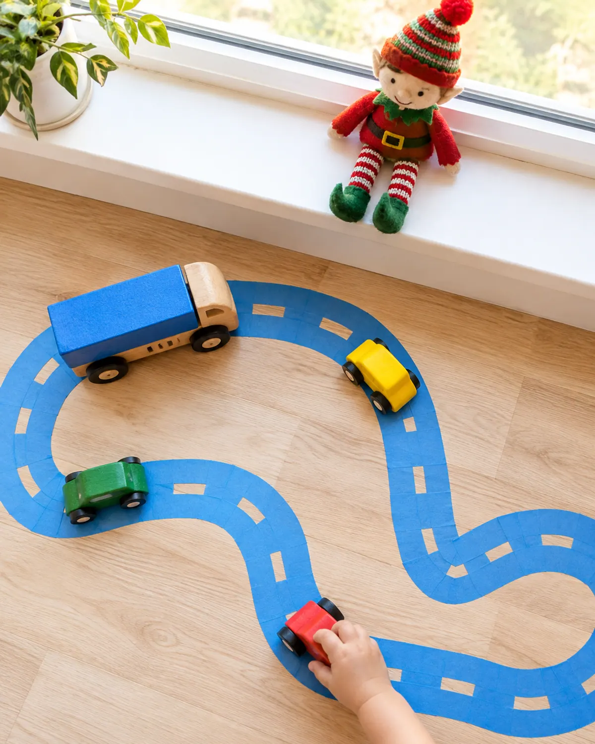 Toddler-safe elf idea: a painter's tape road on a wood floor with chunky toy cars, a small hand pushing a car, and a felt elf watching from a windowsill