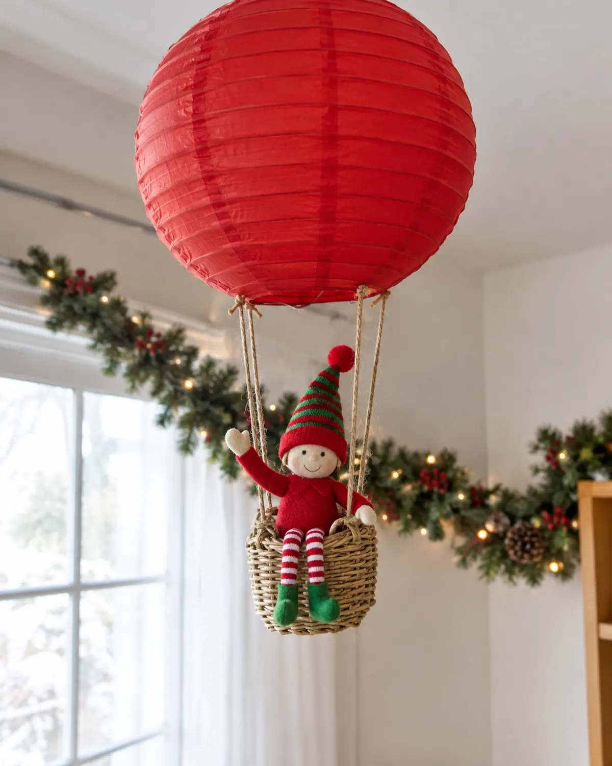 A felt elf doll riding in a small woven berry basket hung under a round red paper lantern like a hot air balloon in a living room