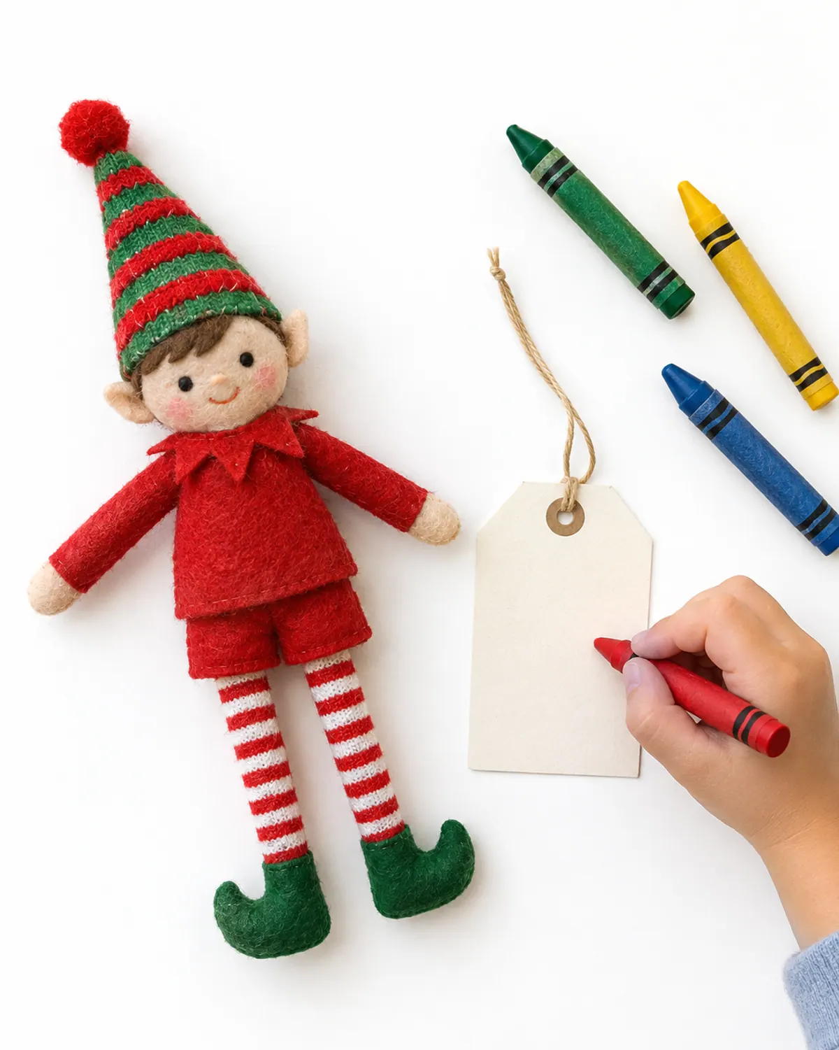 Overhead view of a felt elf doll on a light wood table next to a blank paper name tag, crayons and a small child's hand writing a name