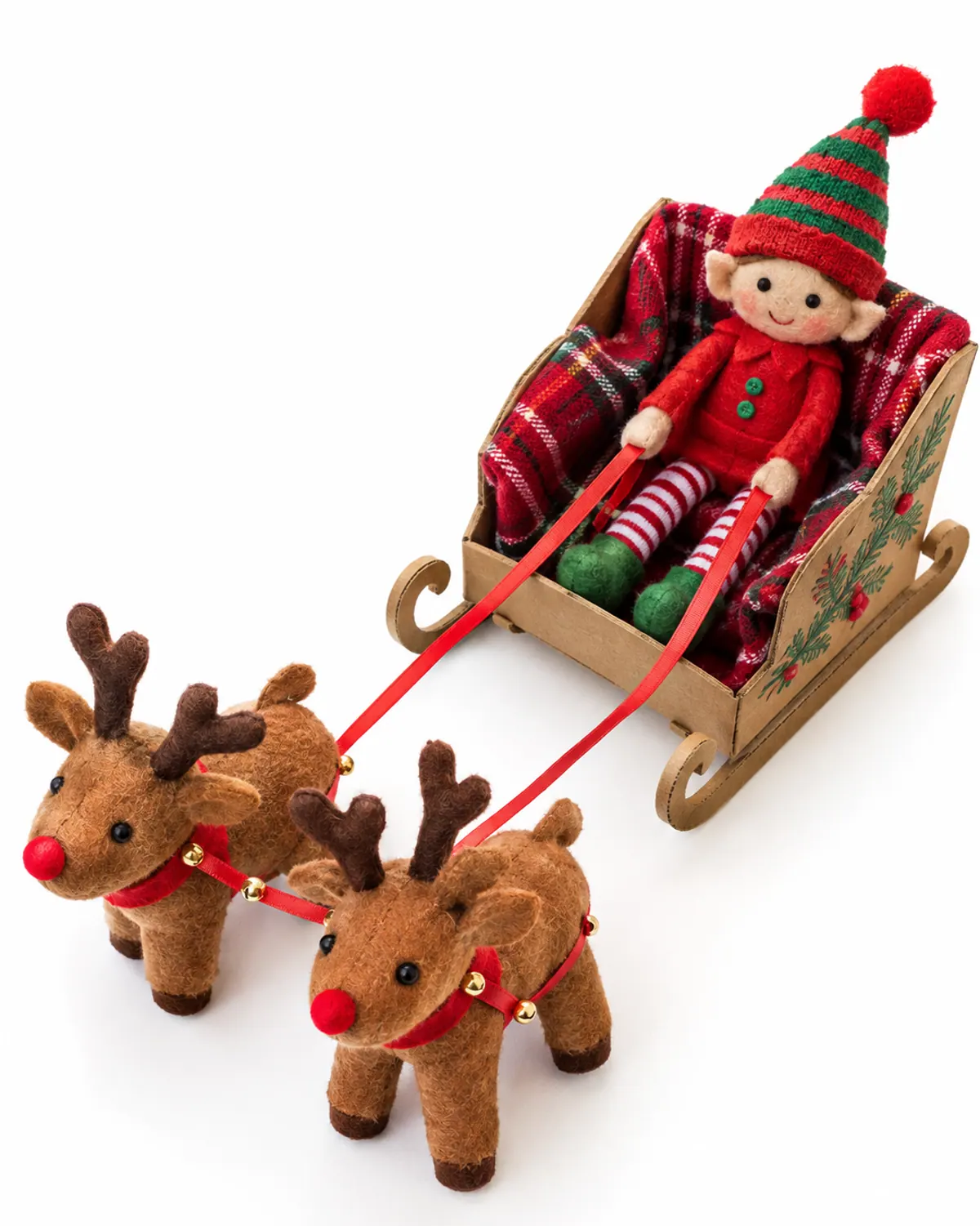 Elf on the shelf arrival: a felt elf doll in a shoebox sleigh lined with a plaid towel, pulled by two plush reindeer toys on a wood floor near a Christmas tree