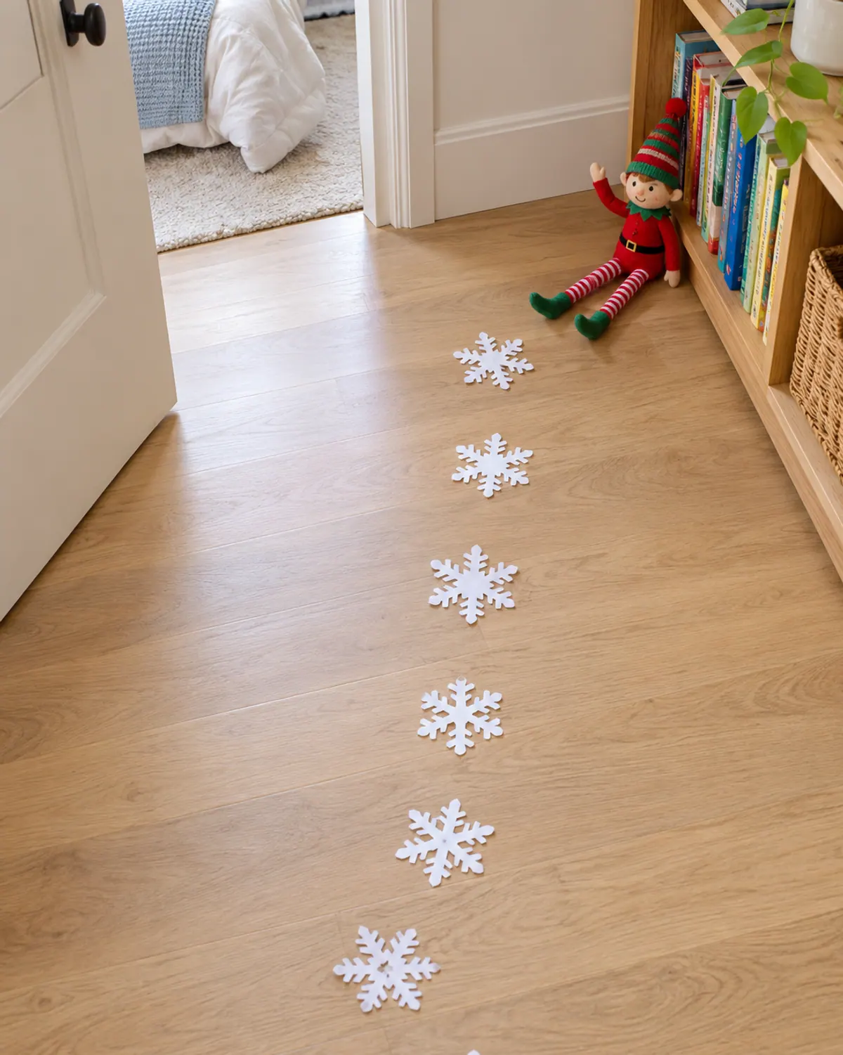 Zero-budget elf arrival: a trail of white paper snowflakes on a wood hallway floor leading to a felt elf doll on a low bookshelf