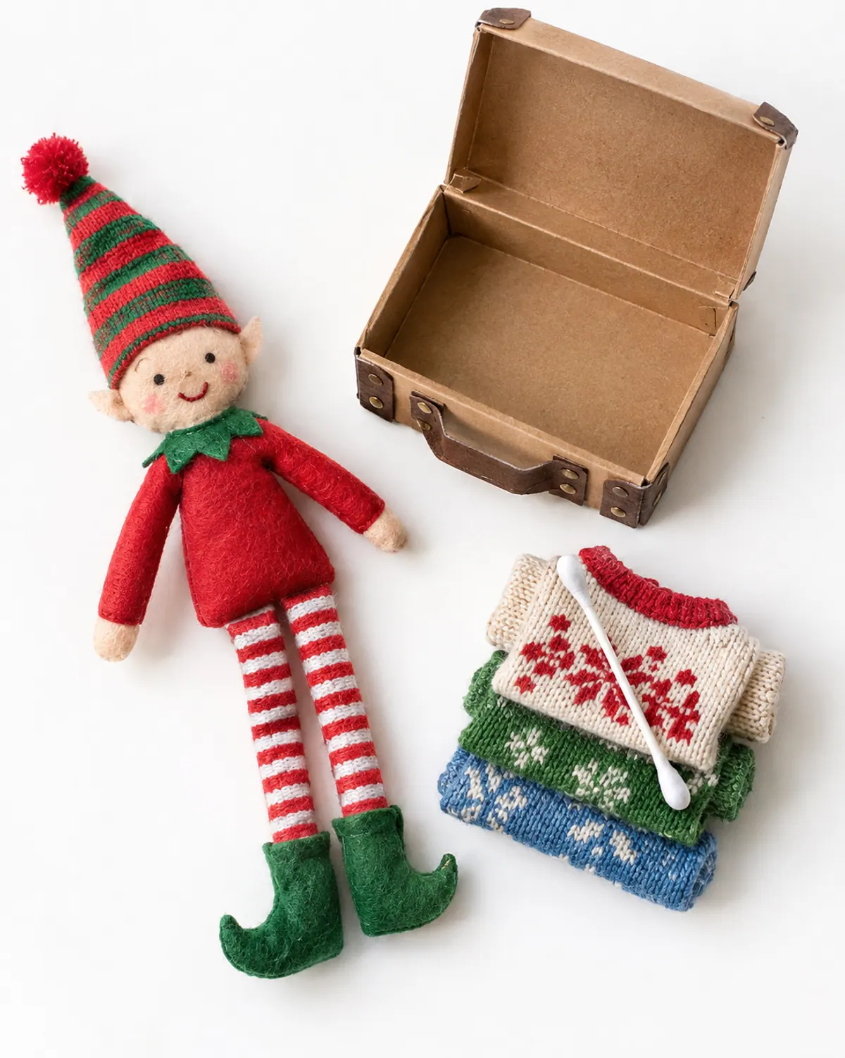 A felt elf doll sitting on a bedroom shelf beside a small open box suitcase with tiny folded doll sweaters and a cotton swab toothbrush