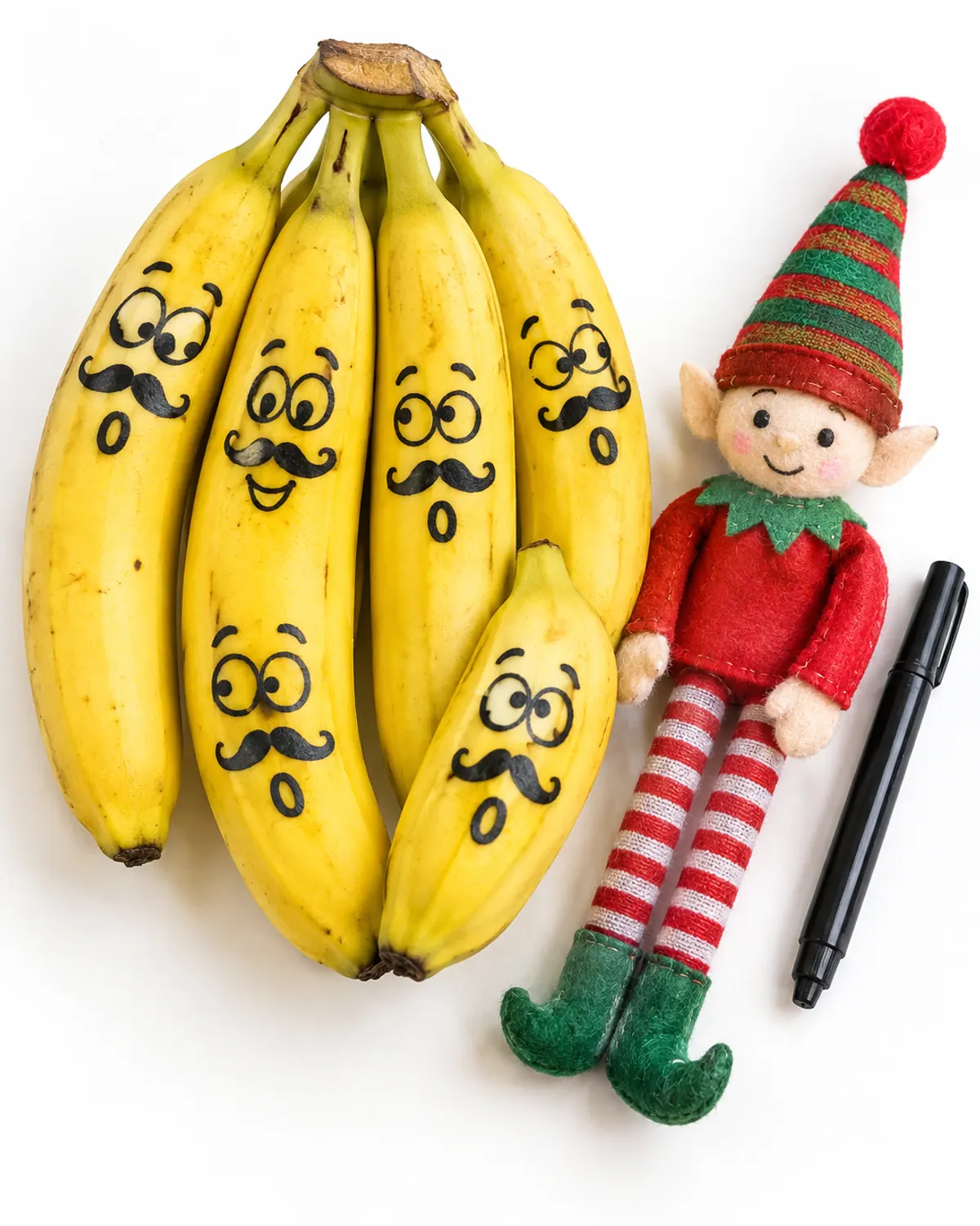 A red felt elf doll holding a black marker next to a bunch of bananas with silly doodled smiles and mustaches drawn on the peels