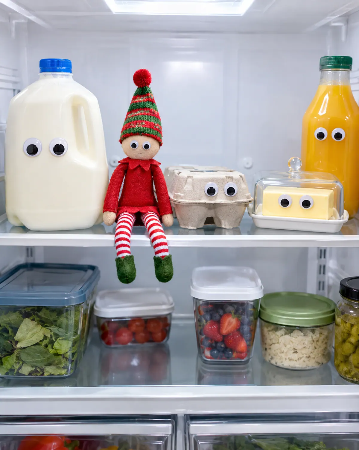 Open fridge with silly googly stickers on the milk jug, eggs and butter dish, and a red felt elf doll sitting on the top shelf