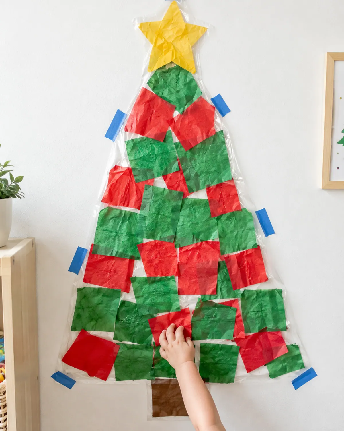 Toddler Christmas craft: a clear contact paper tree taped to a wall with green and red tissue paper squares stuck on, chubby toddler hands pressing on another square