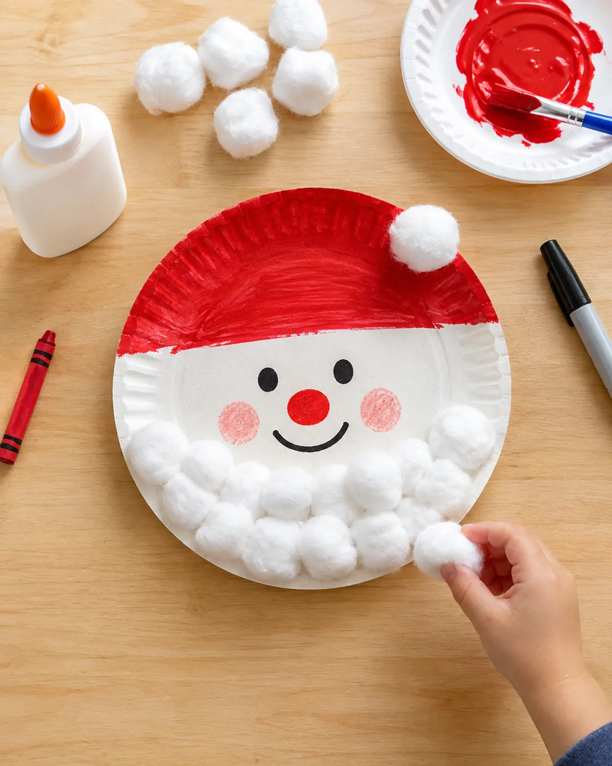 Preschool Christmas craft: a paper plate Santa with a red painted hat and a fluffy cotton ball beard, a preschooler's small hand gluing on one more cotton ball