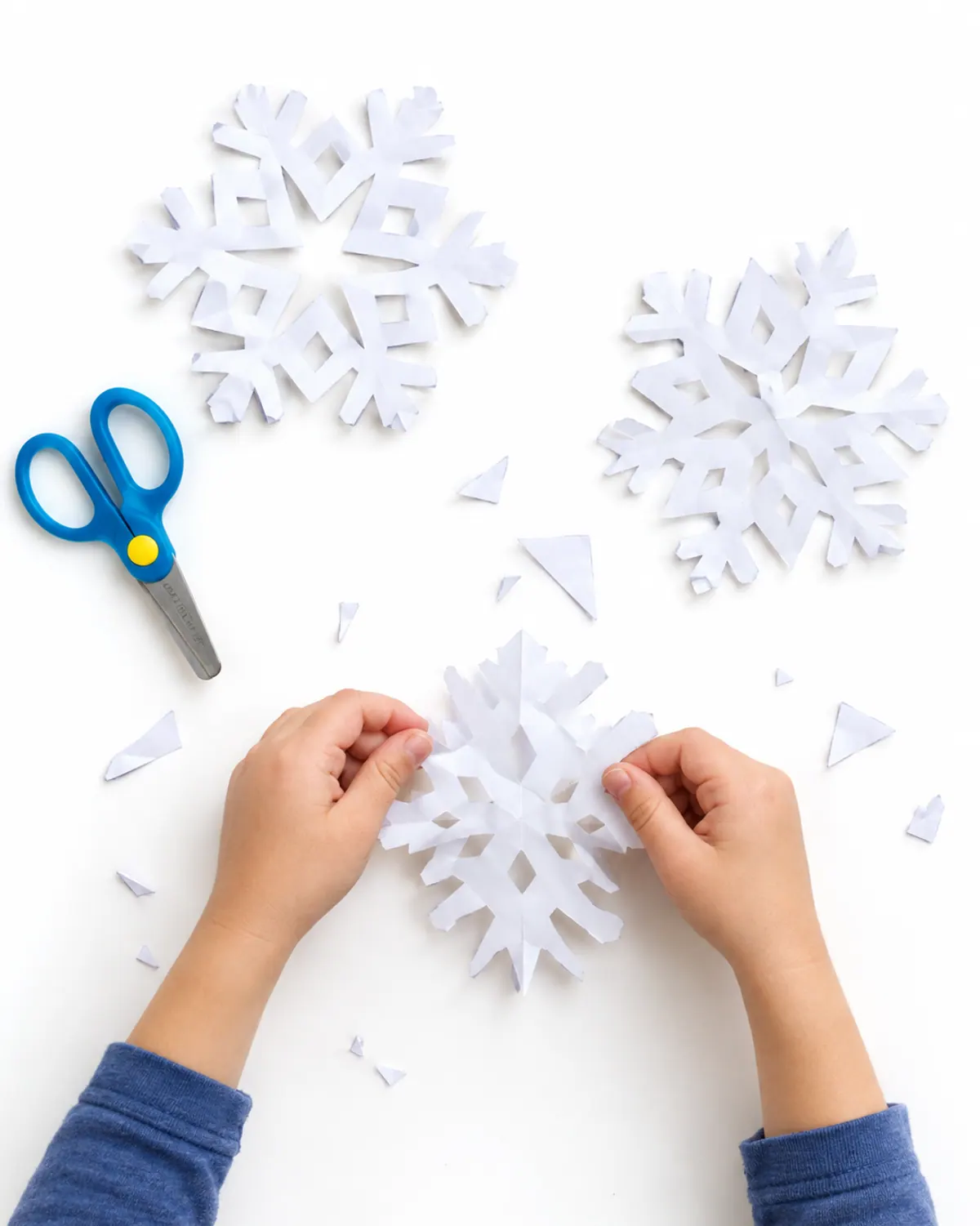 A child's hands unfolding a white paper snowflake on a wood table covered in paper scraps and child-safety scissors