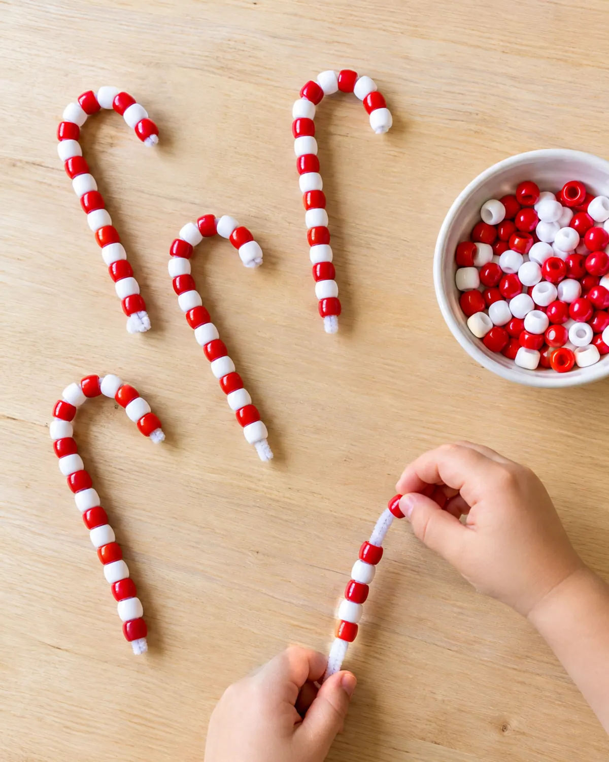Pipe cleaner candy cane ornaments with red and white pony beads on a wood table, a preschooler's hand threading another bead