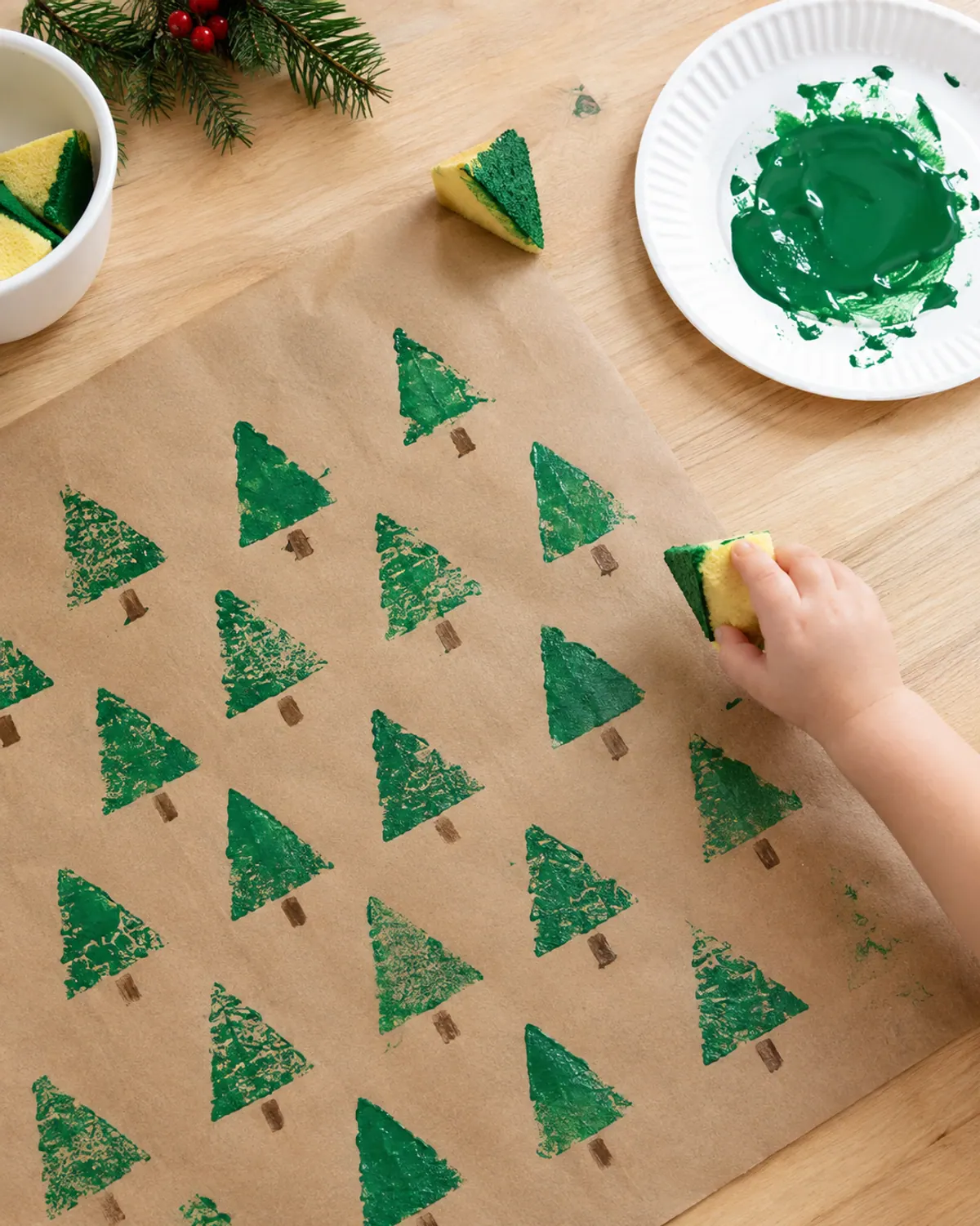 Easy Christmas craft for kids: green triangle sponge stamps making a row of Christmas trees on brown paper, a toddler's small hand holding the sponge