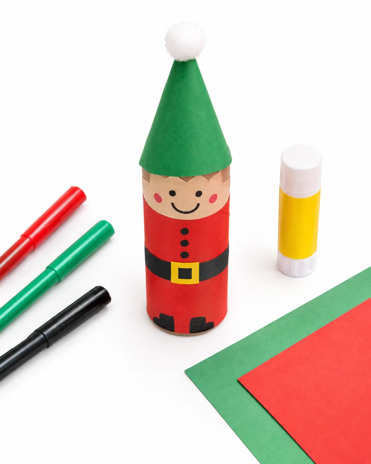 A toilet paper roll elf craft wrapped in red paper with a green paper hat and cotton pom on top, standing next to markers