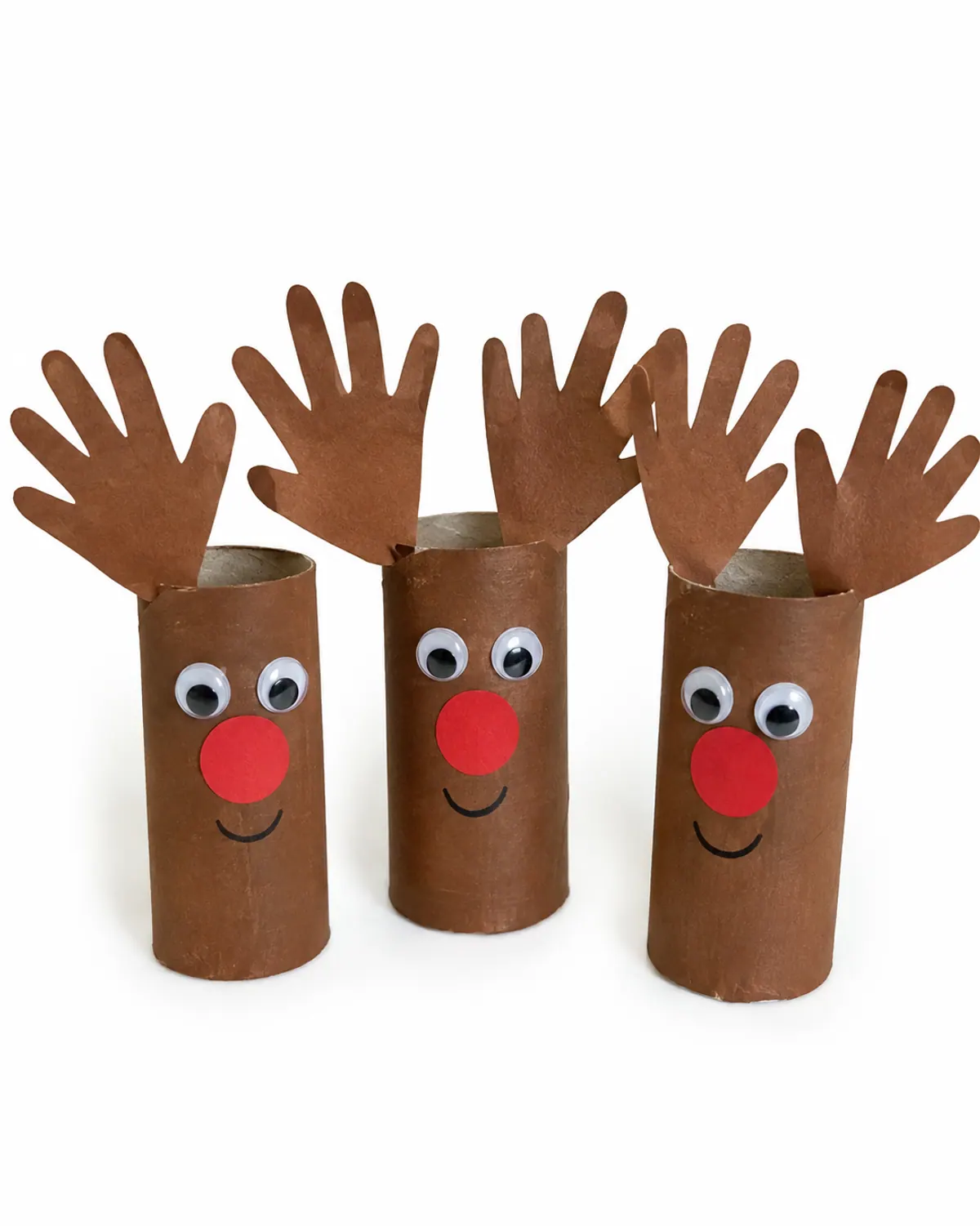 Three toilet paper roll reindeer crafts painted brown with paper handprint antlers and red paper noses standing on a wood table