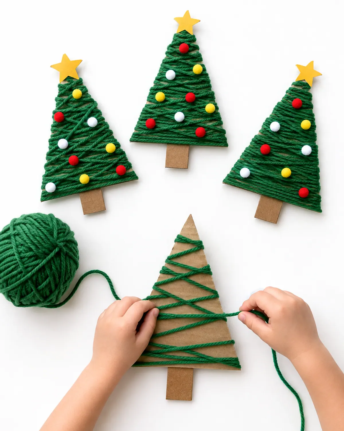 Three cardboard Christmas trees wrapped in green yarn with a child's hands wrapping another on a wood table