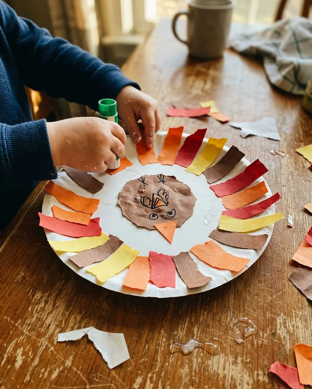 Easy Thanksgiving craft for kids: a paper plate turkey with torn orange, red, yellow and brown paper feathers and marker details, small hands gluing on one more strip